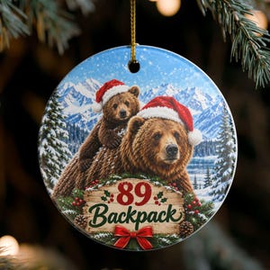 Può includere: Un ornamento rotondo in ceramica con due orsi bruni che indossano cappelli da Babbo Natale su uno sfondo di montagne innevate. Un cartello in legno mostra il testo rosso 89 e il testo verde Backpack, decorato con agrifoglio e un fiocco rosso.