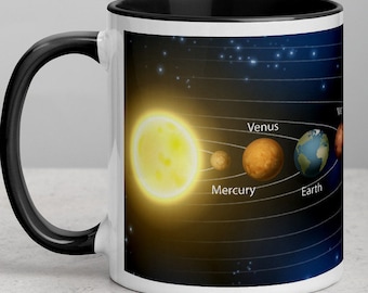 May include: A white coffee mug with a black rim and handle. The mug features a graphic of the solar system with the sun, Mercury, Venus, Earth, and Mars. The planets are labeled with their names.