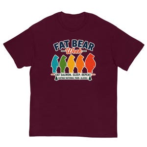May include: A maroon short-sleeved t-shirt featuring a graphic with the text FAT BEAR Week and EAT SALMON. SLEEP. REPEAT. KATMAI NATIONAL PARK, ALASKA. The design includes five colourful bear silhouettes in blue, green, yellow, orange, and red.