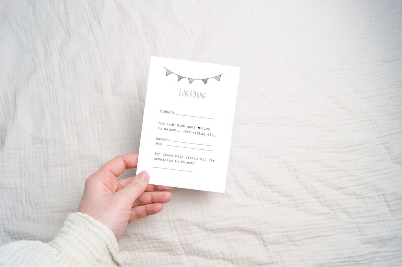 May include: A white card with black text that reads "Einladung" (Invitation) with a string of gray and white triangle flags at the top. The text on the card reads: "Liebe/r, Ich lade Dich ganz herzlich zu meinem Geburtstag ein. Wann?  Wo? Ich freue mich riesig mit Dir gemeinsam zu feiern!"