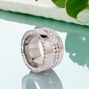 Op de afbeelding: Een zilveren ring met een brede band en twee rijen met heldere zirkonia-stenen. De ring is gegraveerd met "STAINLESS STEEL #204".