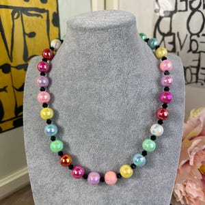 Peut inclure: Un collier de perles colorées avec des perles rondes et irisées de différentes couleurs, dont le rouge, le rose, le jaune, le vert et le bleu. Les perles sont entrecoupées de petites perles noires, créant un accessoire vibrant.