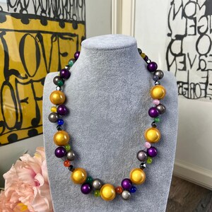 May include: A colorful beaded necklace with large, round, gold-colored beads interspersed with smaller purple, gray, and green beads. The necklace is displayed on a gray mannequin bust, with artwork in the background.