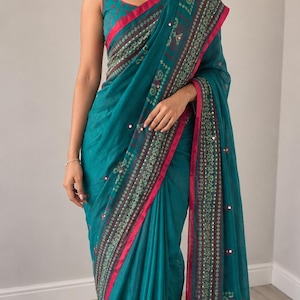 May include: A teal sari featuring a sleeveless blouse with a square neckline, adorned with a dark border, intricate green and white floral patterns, and small reflective mirror embellishments along the fabric edges and throughout the main body.