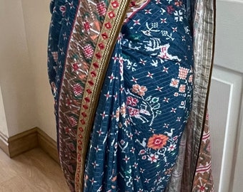 May include: A blue and brown patterned saree with a gold and red trim. The saree is draped over a mannequin and has a tassel on the end.