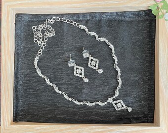 May include: A silver necklace and earrings set with a delicate chain and a pendant with a square design featuring clear rhinestones.