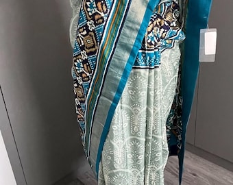 May include: A teal and white patterned saree with a decorative border. The saree is draped over a mannequin and has a matching teal fabric on the floor.