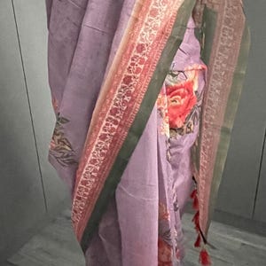 Puede incluir: Un sari lavanda con un estampado floral con grandes flores rojas y naranjas. El sari tiene un borde decorativo con un diseño rojo y dorado y un ribete verde. La tela está drapeada sobre un maniquí.