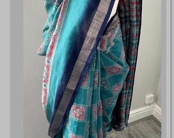 May include: A teal saree with a geometric and floral pattern. The saree features a dark blue border with a silver stripe. The fabric is draped over a mannequin, showcasing the intricate design and color variations.