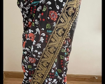 May include: A black saree with a floral print and gold embroidery. The saree has a white border with gold trim. A pearl necklace with colorful beads is draped over the saree.