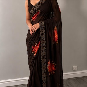 May include: A dark brown saree with a floral design in red and orange. The saree features a decorative gold border. The fabric is lightweight and flowing, ideal for formal events. The saree is a traditional garment.