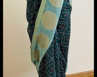 May include: A teal blue saree with a gold and silver patterned border. The saree has a floral design and is draped on a mannequin. The mannequin is wearing a pearl necklace with green and blue accents.