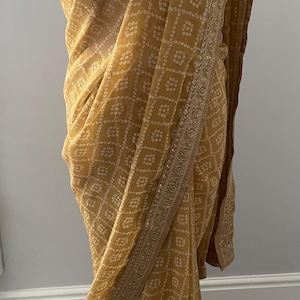 Puede incluir: Sari amarillo mostaza con un patrón de puntos blancos repetidos. El sari tiene un borde decorativo con un diseño detallado. La tela está drapeada sobre una forma, mostrando la textura y el flujo del material.