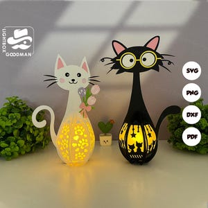 Op de afbeelding: Twee katvormige decoratieve lampen. Eén is wit met een boeket, de andere zwart met een bril. Beide hebben verlichte, patroonbuiken. De lampen zijn gemaakt van uitgesneden materiaal, met het woord "GOODMAN" zichtbaar.