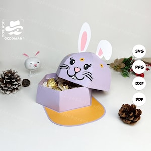 May include: A lilac-colored paper box shaped like a baseball cap, decorated with a bunny face and ears. The box is open, revealing gold-wrapped chocolates. The brim is yellow. A small bunny figurine and pinecone are in the background.