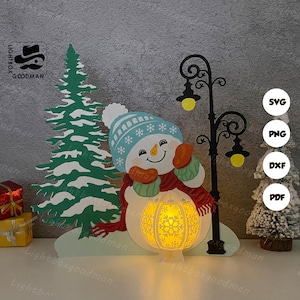 May include: A festive Christmas papercraft scene featuring a snowman with a blue hat and scarf, a green Christmas tree, and a black lamppost. The snowman holds a glowing lantern. The scene is set on a white surface.
