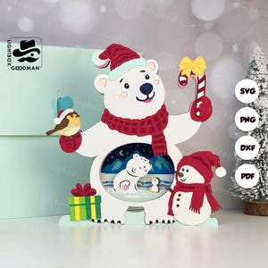 Könnte beinhalten: Weihnachtsdekoration mit einem weißen Eisbären mit rotem Schal und Hut, der eine Zuckerstange hält. Ein Schneemann und ein Vogel sind ebenfalls abgebildet. Die Dekoration hat eine ausgeschnittene Szene eines Eisbären und eines Pinguins.