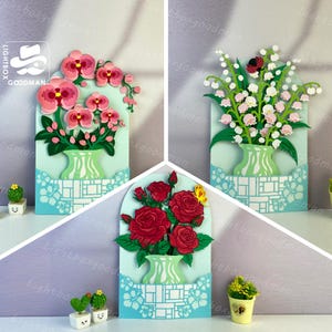 Op de afbeelding: Drie papieren bloemstukken in vazen. De arrangementen tonen roze orchideeën, witte lelietjes-van-dalen en rode rozen. Elk arrangement staat tegen een lichtblauwe achtergrond met een decoratieve rand. Het Goodman Lightbox-logo staat linksboven.