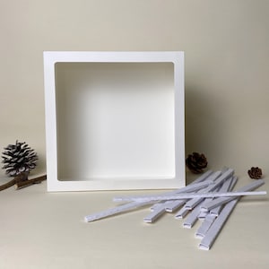 May include: A white, square shadow box with a recessed interior, suitable for displaying art or collectibles. Several white, rectangular mounting strips are in front of the box. Pinecones and a twig are in the background.