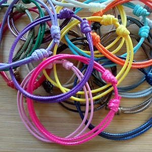 May include: A selection of brightly coloured cord bracelets, knotted and piled together. The bracelets are in shades of pink, purple, yellow, and blue. The knots and cords are clearly visible. The background is a light brown wooden surface.