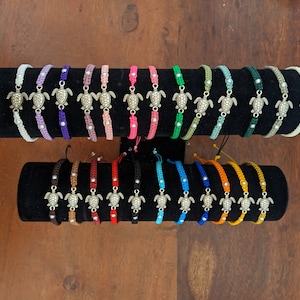 May include: A display of colourful string bracelets with silver turtle charms. The bracelets are arranged in rows on a black display stand.