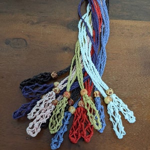 May include: A selection of colourful macrame plant hangers in shades of blue, red, green, and pink. Each hanger has a woven net design and a wooden bead. The hangers are displayed on a dark wooden surface.