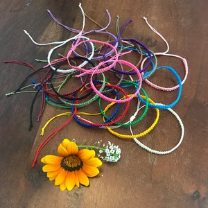 May include: A collection of colorful friendship bracelets made with knotted string. The bracelets are arranged in a circle on a wooden surface with a yellow flower in the center.
