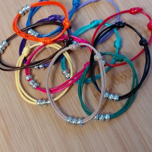 May include: A selection of colourful bracelets with adjustable knots and silver-coloured beads. The bracelets are in various colours, including blue, orange, pink, yellow, green, and black. The bracelets are displayed on a wooden surface.