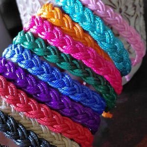 May include: A selection of braided bracelets in a spectrum of colours: pink, orange, teal, green, blue, purple, red, beige, and black. The image is a close-up, highlighting the woven texture and vivid shades. The bracelets are likely handmade, and are a variety of sizes.