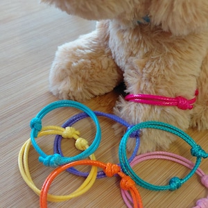 May include: A selection of brightly coloured bracelets in orange, yellow, blue, purple, and pink. The bracelets are made of cord and knotted. A plush dog toy is in the background.
