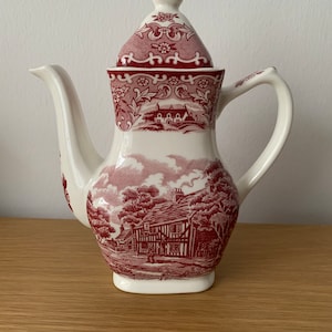 May include: A white ceramic teapot with a red and white floral design. The teapot has a lid and a spout. The design features a small village scene.