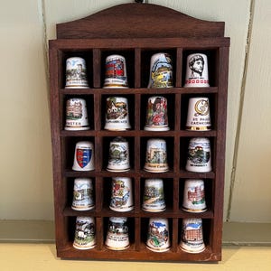May include: A dark brown wooden wall-mounted display case with twenty square compartments, each holding a white ceramic thimble. Each thimble features a unique colorful illustration and text depicting various landmarks, buildings, and historical sites.