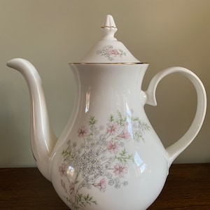 May include: White porcelain teapot with a floral design featuring pink and grey flowers. The teapot has a gold trim around the lid and a gold band around the base.