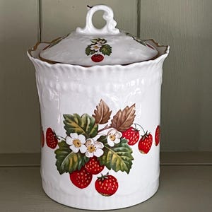 May include: A white ceramic canister with a lid featuring a decorative handle. The body and lid are adorned with a colourful pattern of red strawberries, green leaves, and white blossoms. The rim of the lid has a gold-coloured trim detail.