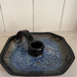 May include: A ceramic candle holder with a black handle and a dark blue interior. The holder has a hexagonal shape with a dark brown and black rim. The interior has a speckled pattern.