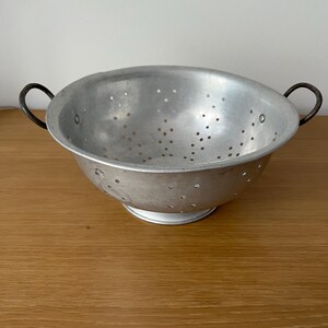 May include: A silver colander with two black handles. The colander has a round base and is made of metal.