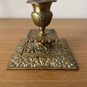 Peut inclure: Un chandelier en laiton vintage avec un design orné. La base est carrée et présente des motifs floraux complexes. Le porte-bougie est une simple coupe ronde.