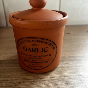 May include: A terracotta garlic storage jar with a matching lid. The front features a black oval label with the text: THE ORIGINAL SUFFOLK CELLAR, GARLIC, Ideal Storage at Home or on Safari, MADE BY HENRY WATSON POTTERY ENGLAND, Founded 1800.