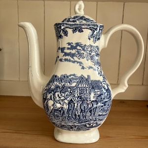 May include: A white and blue ceramic coffee pot with a lid. The pot features a detailed blue transferware design depicting a rural scene with horses and figures. The handle and spout are white.