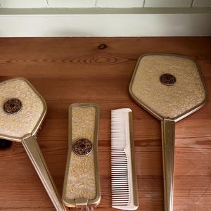 May include: A vanity set consisting of a hand mirror, a hairbrush, and a comb. Each piece features a gold-tone metal frame with a textured gold floral fabric inlay and a central circular medallion containing small amber-colored stones.