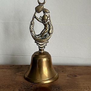 Peut inclure: Une cloche en laiton vintage avec un design détaillé. La cloche présente une petite figurine dans un cadre ovale, avec les mots "Cornish Pixie" inscrits en dessous. La cloche a une boucle en haut pour l'accrocher et repose sur une surface en bois.