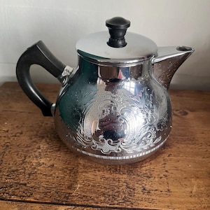 May include: A silver-colored teapot with a black handle and lid. The teapot has an ornate floral design etched into the metal. The spout is curved, and the lid has a black knob. The teapot is sitting on a wooden surface.
