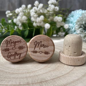 May include: Wooden bottle stoppers with engraved text and arrow designs. One stopper reads "Morgane Geoffrey", the other "Merci". A third stopper is made of cork and wood. The background features white and blue flowers.