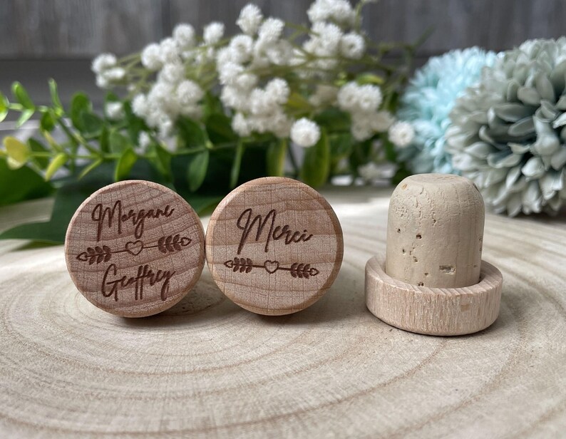 May include: Wooden bottle stoppers with engraved text and arrow designs. One stopper reads "Morgane Geoffrey", the other "Merci". A third stopper is made of cork and wood. The background features white and blue flowers.