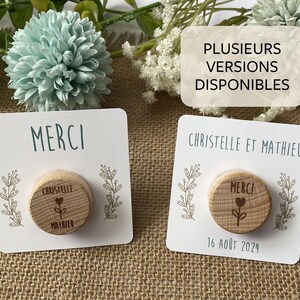 May include: Two wooden round magnets with the word "MERCI" and a floral design on a white card. One magnet has the names "Christelle" and "Mathieu" and the date "16 ao&ucirc;t 2024". The background includes flowers.