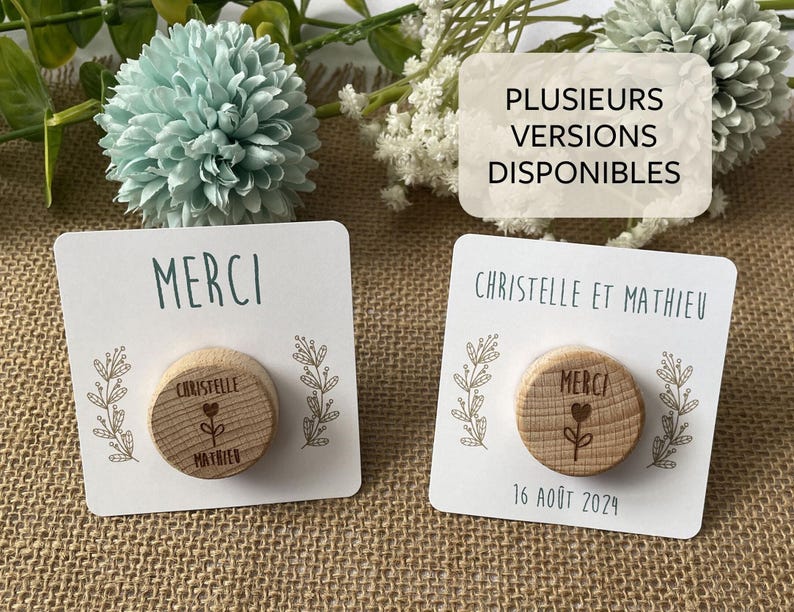 May include: Two wooden round magnets with the word "MERCI" and a floral design on a white card. One magnet has the names "Christelle" and "Mathieu" and the date "16 ao&ucirc;t 2024". The background includes flowers.