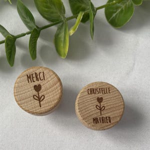 May include: Two small, round wooden objects with engraved text and a floral design. One reads "MERCI" above a heart and flower. The other reads "CHRISTELLE" above a heart and flower, with "MATHIEU" below.
