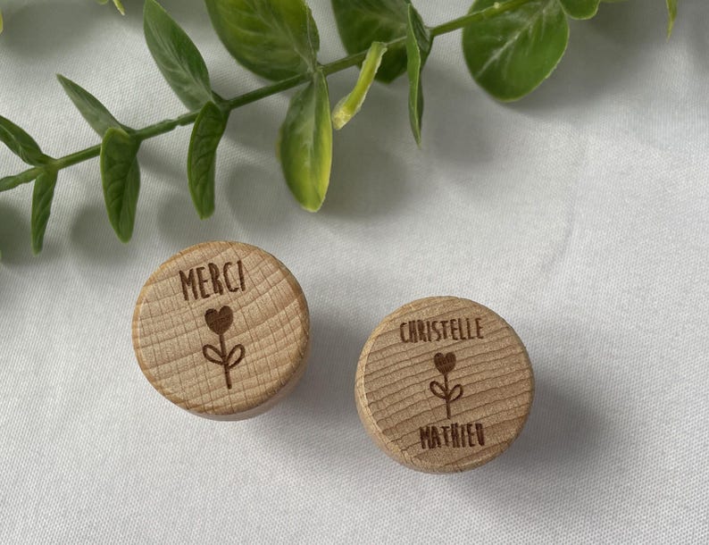 May include: Two small, round wooden objects with engraved text and a floral design. One reads "MERCI" above a heart and flower. The other reads "CHRISTELLE" above a heart and flower, with "MATHIEU" below.