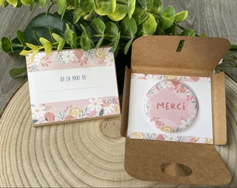 Op de afbeelding: Een cadeau set met een bloemknop met de tekst "Merci" in roze, en een bijpassende kaart met de tekst "De la part de". De set wordt gepresenteerd in een bruine kartonnen doos, met een houten achtergrond.