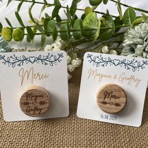 May include: Two white cards with a floral design and the word "Merci" in gold script. Each card has a small, round wooden magnet with engraved text. One magnet says "Morgane & Geoffrey" and the other says "Merci".